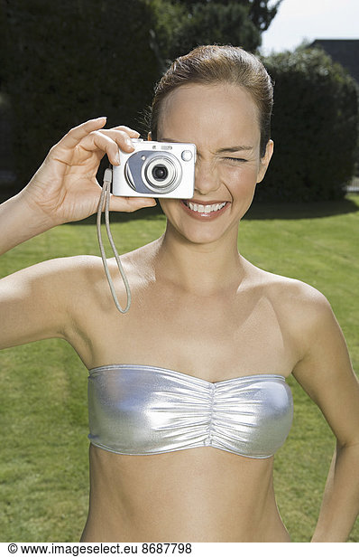 A young woman wearing a silver bandeau top  taking a photograph with a small camera.