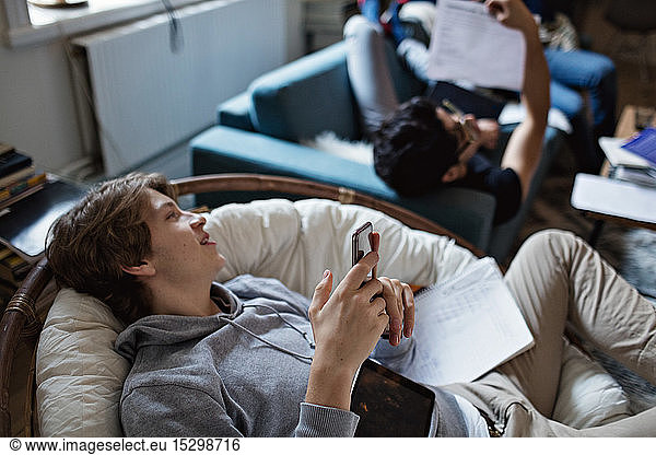 Abgelenkter Teenager nutzt soziale Medien auf dem Handy  während er mit einem Freund zu Hause lernt