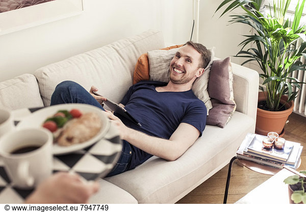 Abgeschnittenes Bild eines jungen schwulen Mannes mit Frühstück für den auf dem Sofa liegenden Partner.