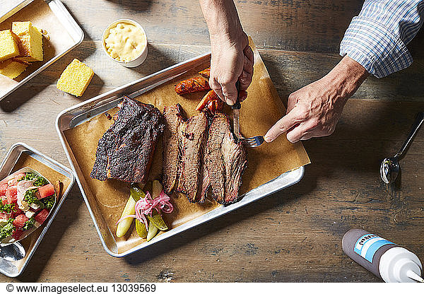 Abgetrennte Hände eines Mannes mit Besteck beim Schneiden von gebratenem Fleisch auf dem Tisch