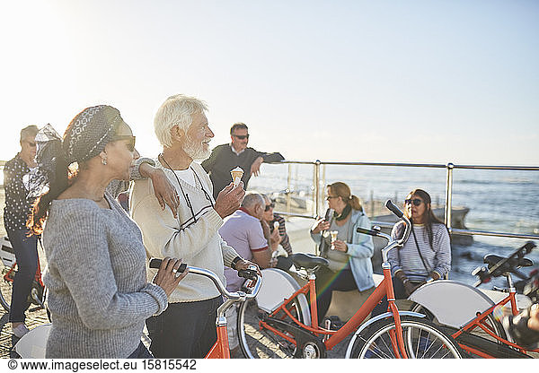 Active senior friend tourists with bicycles eating ice cream at sunny ocean overlook