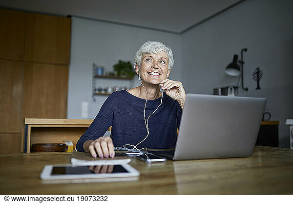 Active senior woman listening to music on smart phone and using laptop while looking away at home
