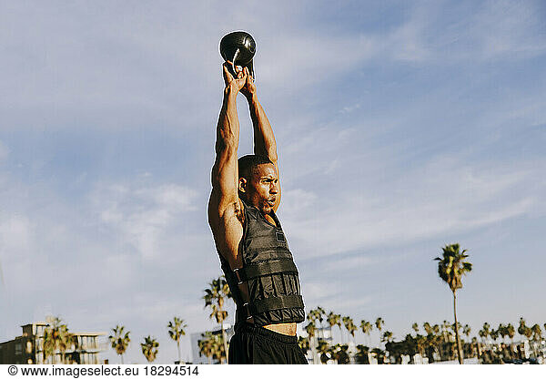 Active sportsman practicing exercise with kettlebell under sky