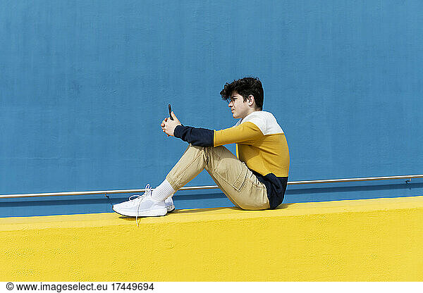 Adolescent sitting on yellow wall using mobile phone against blue wall