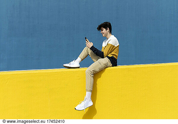 Adolescent sitting on yellow wall using mobile phone against blue wall