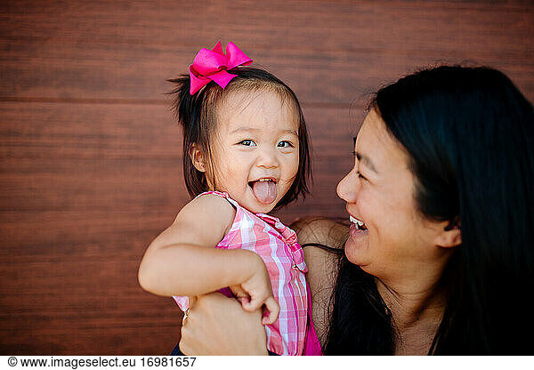 Adorable asian girl held by laughing mom sticks out tongue
