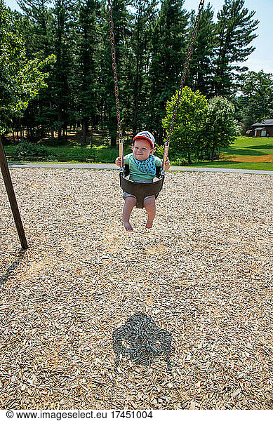Adorable baby boy wearing baseball cap swinging alone at the park.