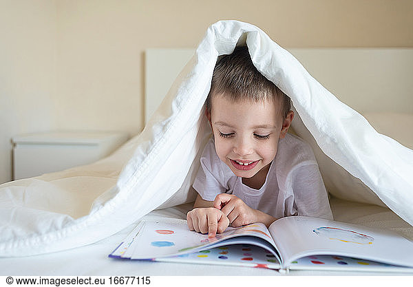 Adorable smiling kid lying down on bed under blanket  reading a book