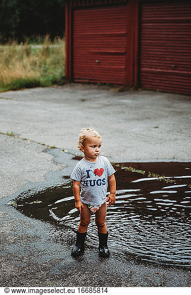 Adorable young boy pouting wearing rubber boots next to a puddle