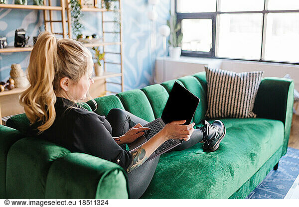 Adult blonde woman lounging on velvet couch holding laptop