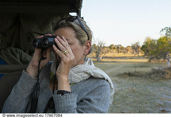 Adult woman looking through binoculars standing by a jeep.