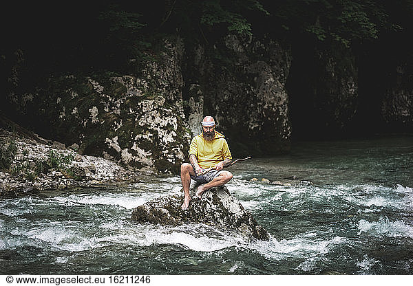 Adventurer with beard sitting in the middle of river on stone and carving on piece of wood