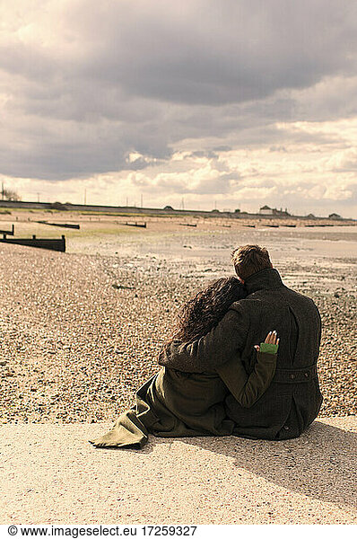 Affectionate couple in winter coats relaxing on sunny tranquil beach