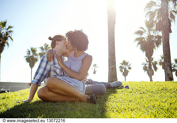 Affectionate couple kissing while sitting on field against sky