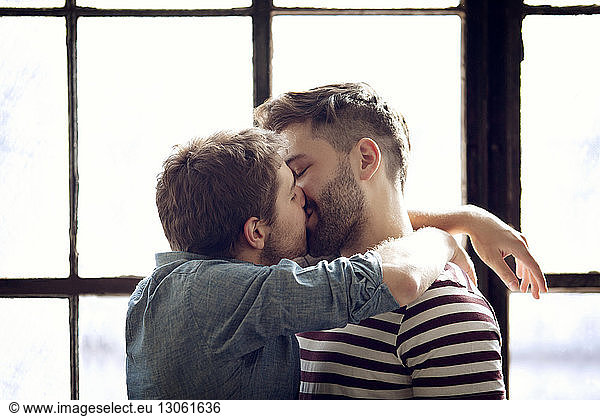 Affectionate gay couple kissing by window at home