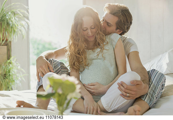 Affectionate pregnant couple kissing in pajamas on bed in sunny bedroom
