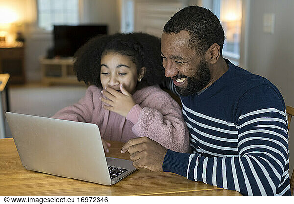 African-American father and tween daughter laugh looking at laptop