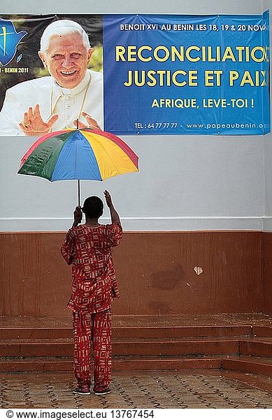 Afrikaner  hinten  trägt einen Regenschirm vor einem Plakat  das den Besuch von Papst Benedikt XVI. in Benin ankündigt.