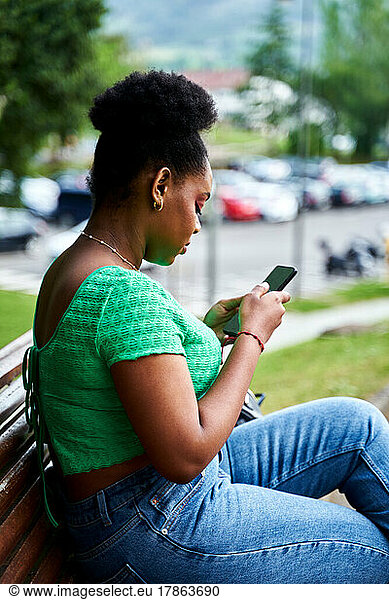 Afro american woman sitting on bench with mobile phone in park