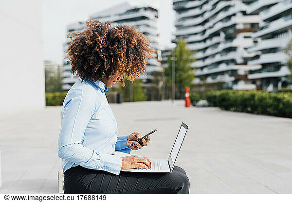 Afro businesswoman using mobile phone sitting with laptop in city