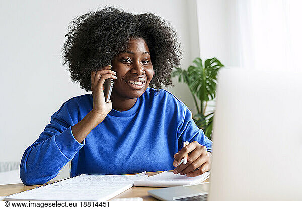 Afro freelancer talking on smart phone in front of laptop at desk