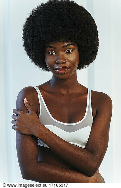 Afro-Model in weißen Dessous umarmt sich  während sie gegen eine weiße Wand steht