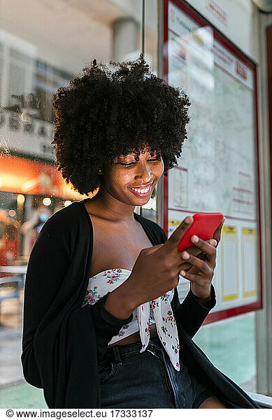 Afro woman smiling while using smart phone at bus stop