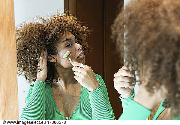 Afro young woman massaging face with jade roller at home