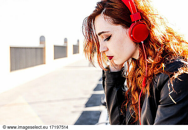 Alternative girl with blue eyes and orange hair listening music with read headphones in an urban space.