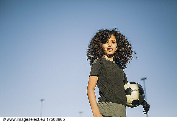 Ambitious girl standing with soccer ball against clear sky