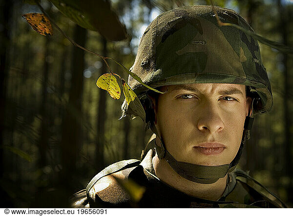An airman stands under a tree during a security forces demonstration. He is wearing a camouflage uniform  body armor and helmet.