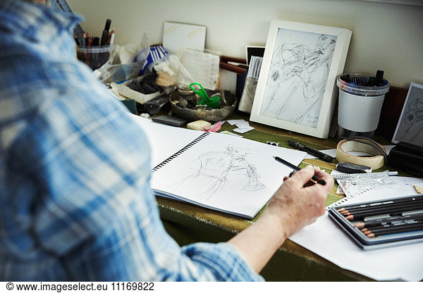 An artist working  holding a pencil over a sketch in progress in an open sketchbook.