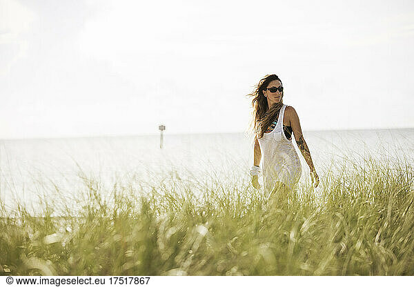 An attractive woman walks along shore with long grass in foreground