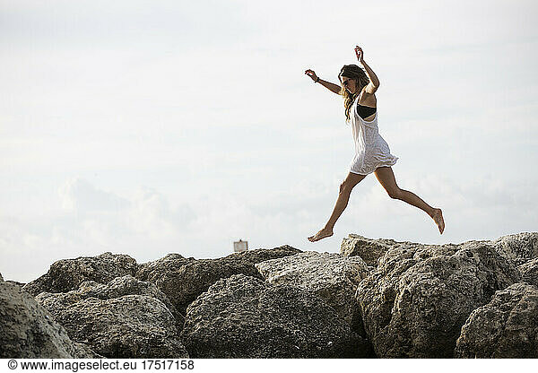 An attractive young woman jumps mid air along shoreline rocks