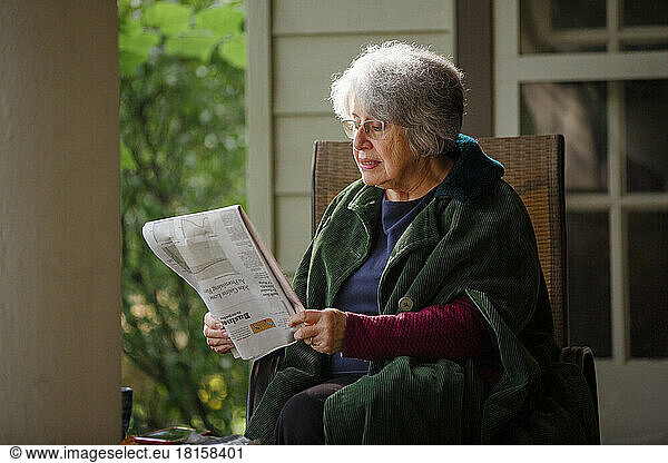 An elderly woman sits on front porch in cloak reading newspaper