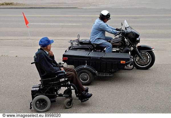 An eldery man in a motorized wheelchair looks at a Harley Davidson mortorcycle speeding by.