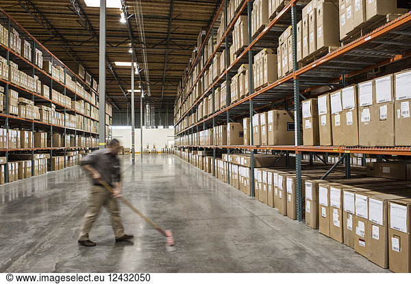 An employee sweeping the floor of an aisle between large racks of storage boxes in a distribution warehouse.