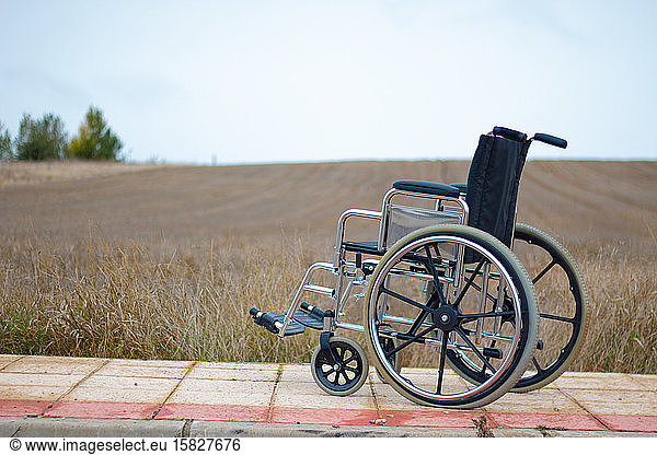 An isolated wheelchair on the sidewalk next to a field.