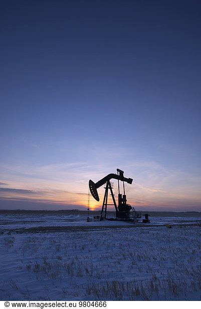 An oil drilling rig and pumpjack on a flat plain in the Canadian oil fields at sunset.