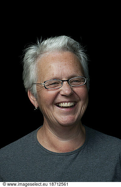 An older Canadian women with short silver hair and designer eyeglasses smiles broadly in a studio built on the sidewalk on the corner of Broadview Avenue and Gerrard Street Avenues