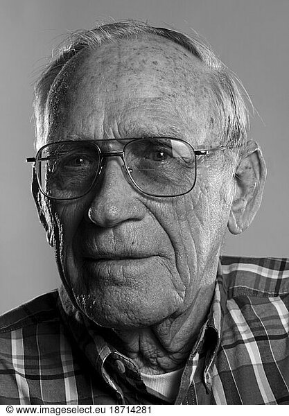 An older veteran man wearing big glasses and a plaid shirt poses for a portrait.