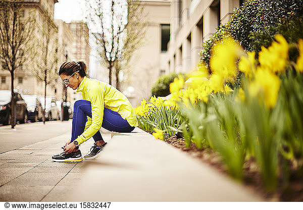 An woman ties her sneakers near some daffodils on a city street.