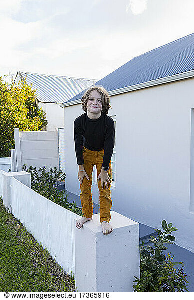 An 8 year old boy standing on a wall outside a house  posing for the camera.