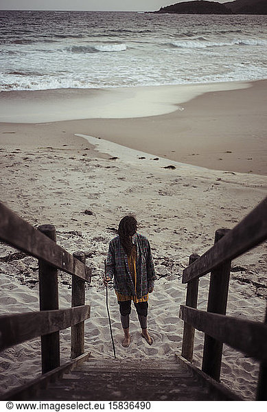 androgynous girl stands at base of beach steps leading to moody ocean