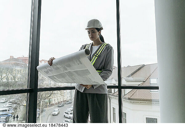 Architect reviewing blueprint standing in front of window at office