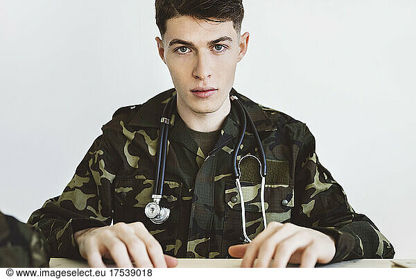 Army doctor with stethoscope sitting in front of white wall