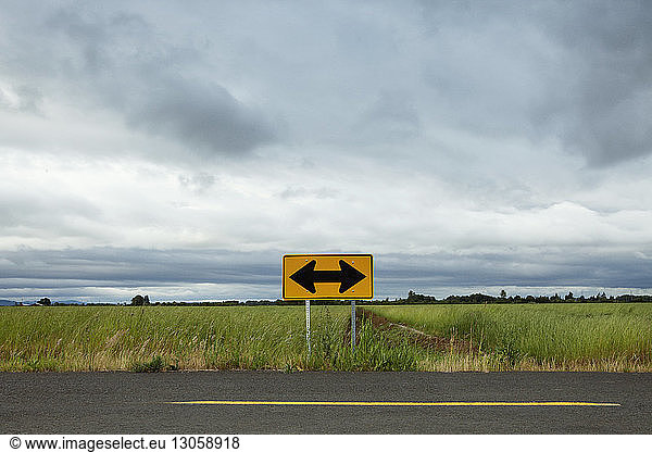 Arrow symbol on empty road by field against cloudy sky