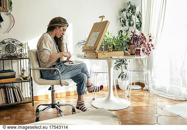 Artist mixing colors in palette sitting on chair in front of easel at home