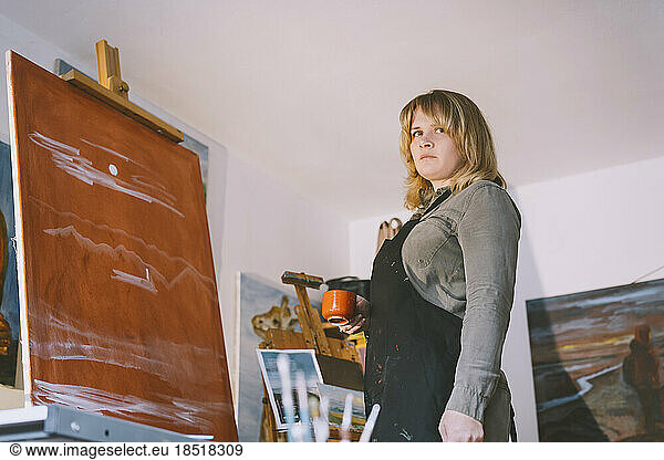 Artist with coffee cup standing in front of canvas at art studio