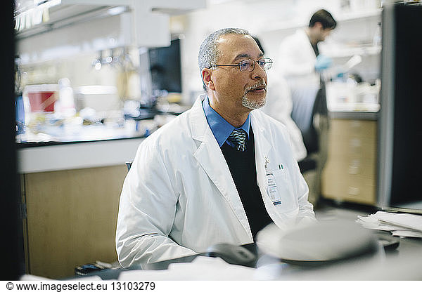 Arzt arbeitet am Schreibtisch  während ein männlicher Kollege im Hintergrund im Krankenhaus arbeitet